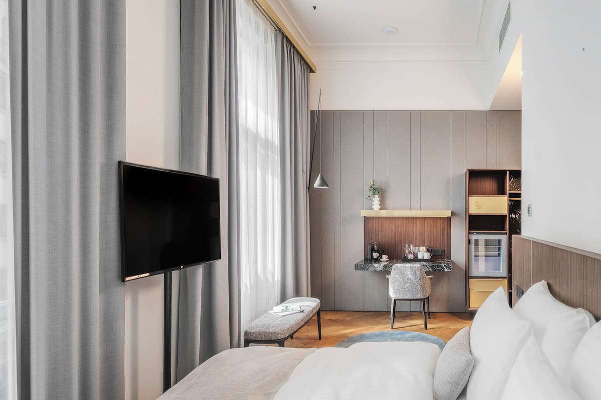 A modern hotel room with a flat-screen TV mounted on the wall, a small seating area with a bench, a kitchenette with a countertop and oven, and a large window with curtains.