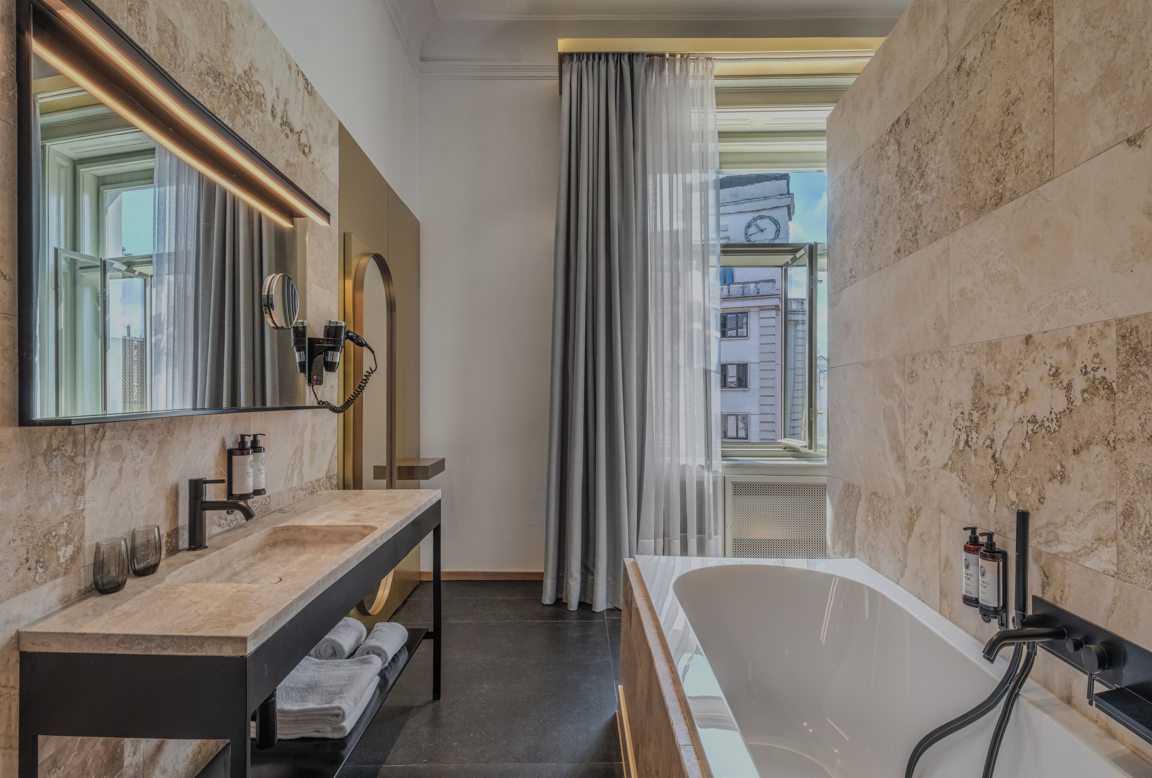A modern bathroom with a large mirror, two sinks, a bathtub, and a view of buildings through a window.
