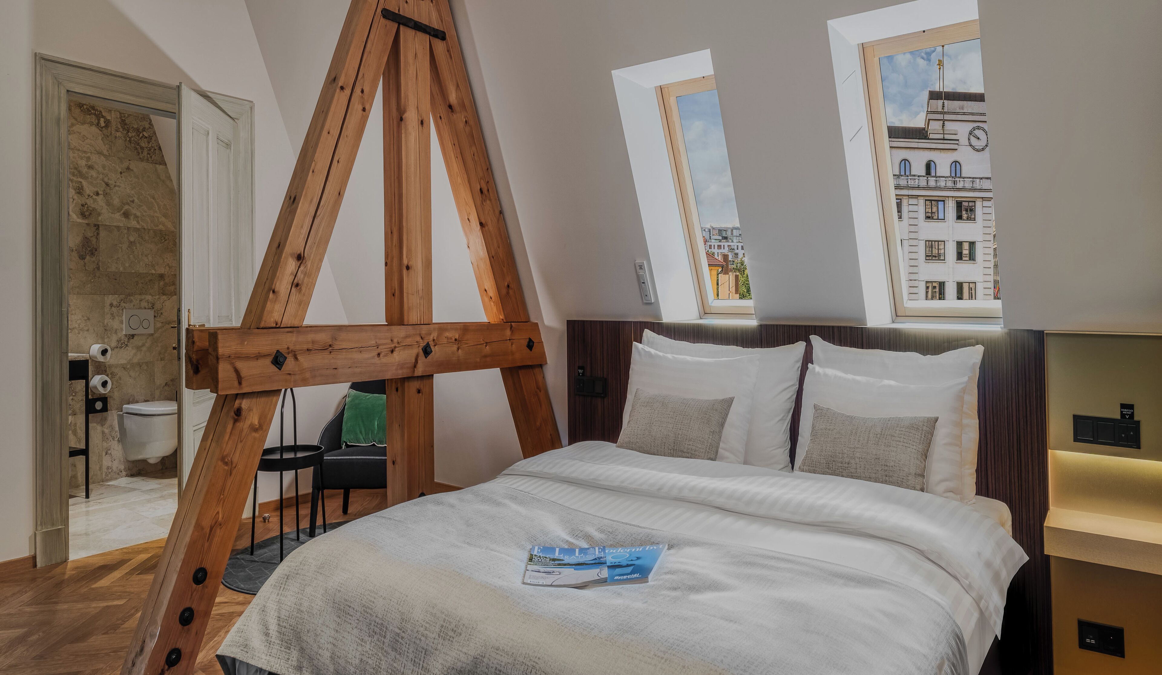 A bedroom with a wooden ceiling structure, a bed with white bedding, a green chair, and a view of buildings through a window.
