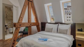 A bedroom with a wooden ceiling structure, a bed with white bedding, a green chair, and a view of buildings through a window.
