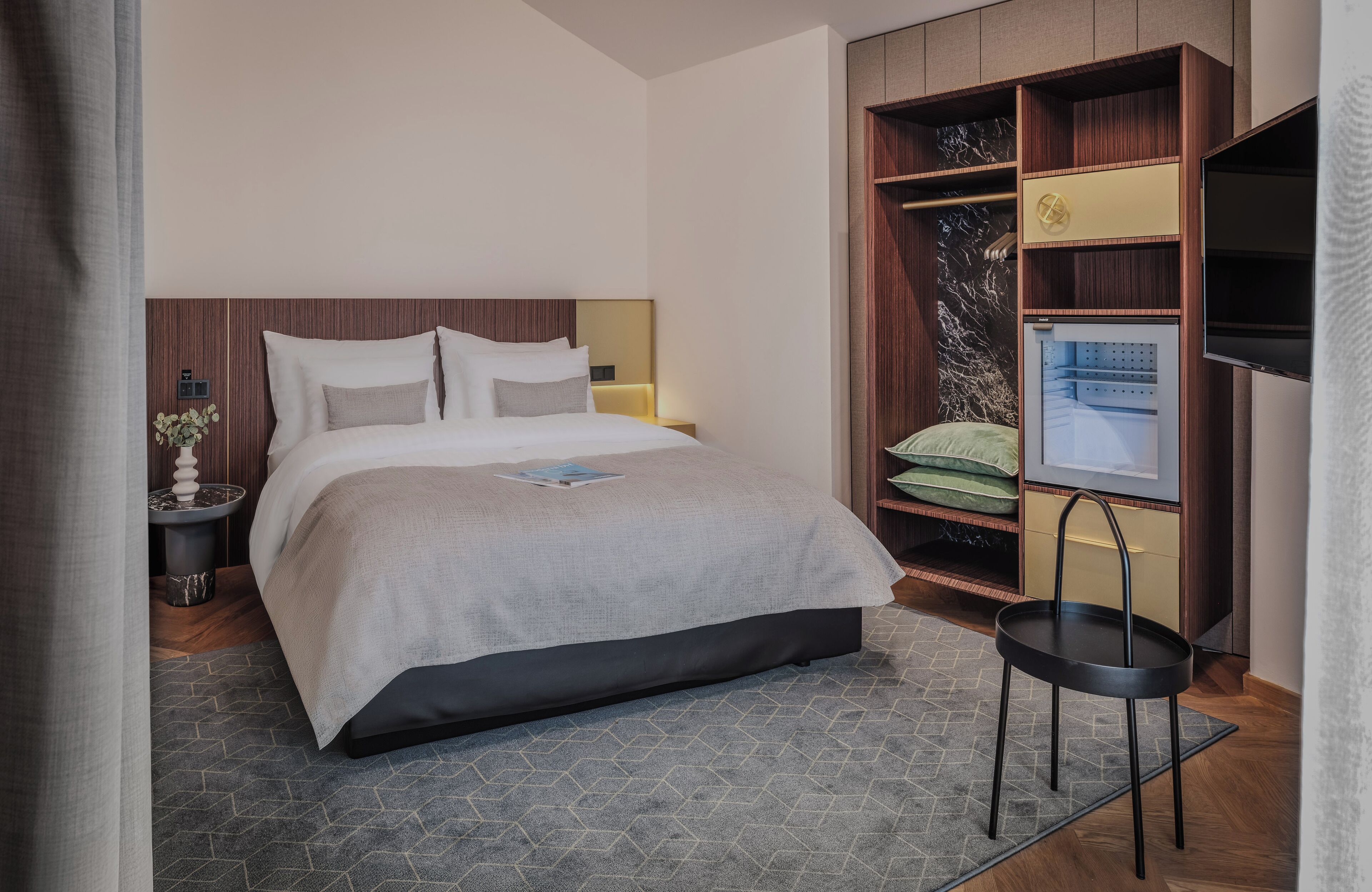 A modern hotel room with a large bed, a small side table, a built-in TV, and a wardrobe with shelves.