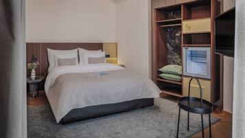 A modern hotel room with a large bed, a small side table, a built-in TV, and a wardrobe with shelves.