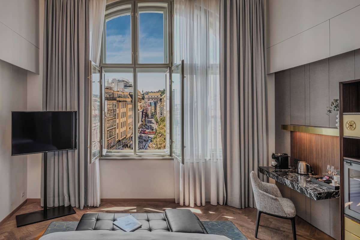 A modern hotel room with a large window, a flat-screen TV, a sofa, a dining area, and a kitchenette.