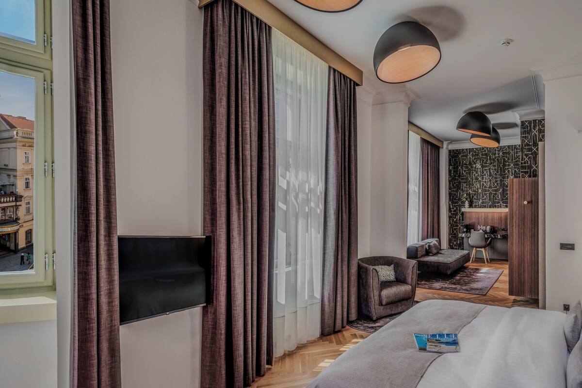 A hotel room with a large bed, a TV, a desk with a chair, and a view of the street.