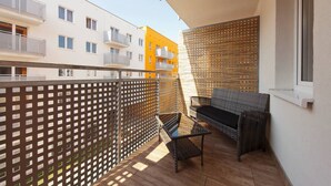 En balkong med en soffa av rotting och ett litet bord, trägolv och utsikt över de intilliggande byggnaderna.