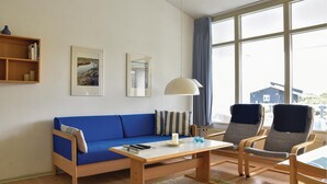 Een moderne woonkamer met een blauwe bank, twee fauteuils, een houten salontafel en een groot raam met uitzicht op een huis.