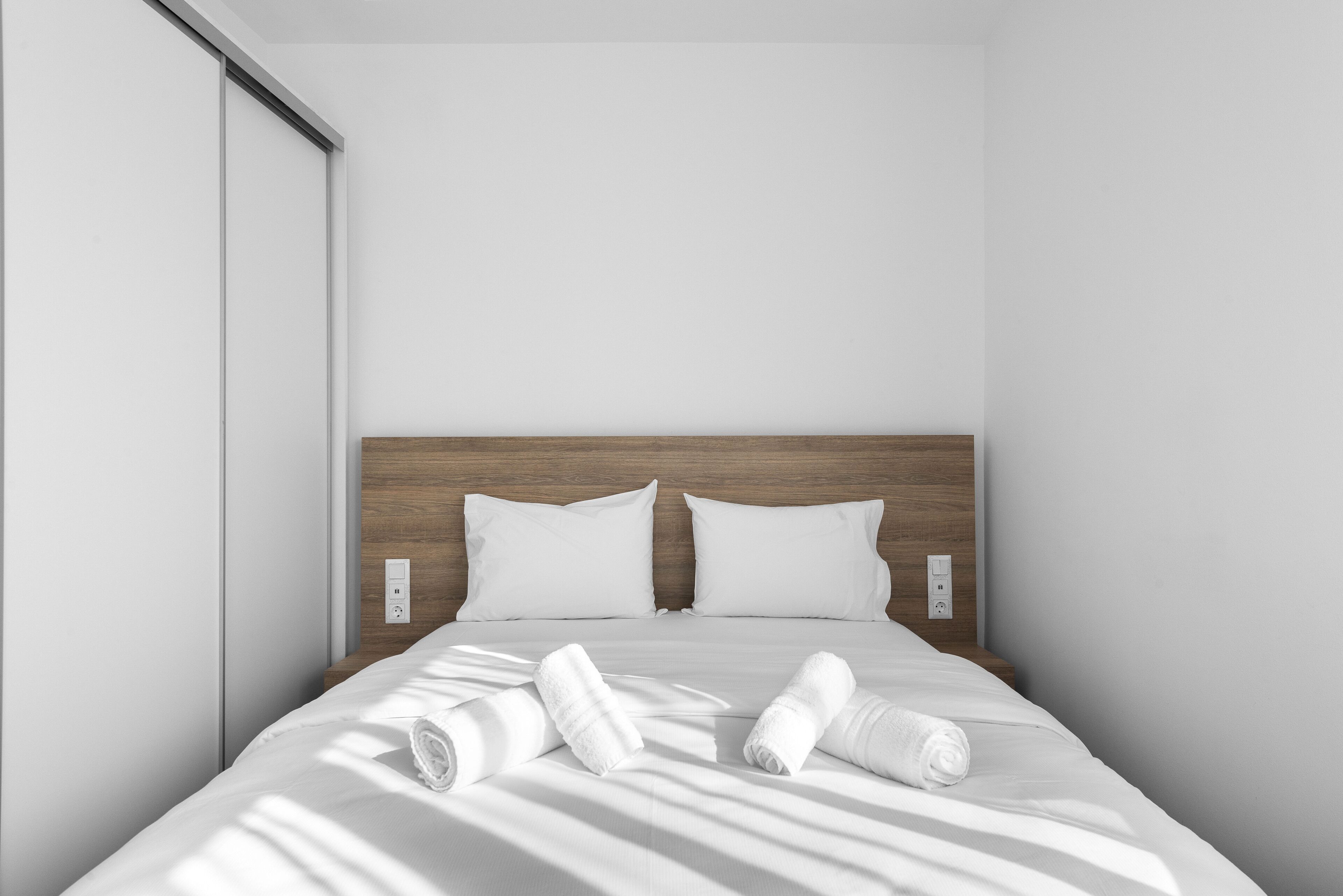 Une chambre d’hôtel moderne avec une tête de lit en bois, du linge de lit blanc et des serviettes soigneusement pliées sur le lit.