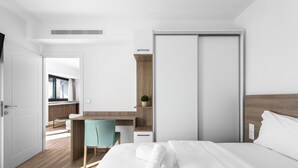 A modern hotel room with a wooden floor, a bed with white linens, a desk with a chair, a wardrobe, and a small plant on the desk. - Penthouse Level 8 Nilie Hospitality MGMT (Thessaloniki)