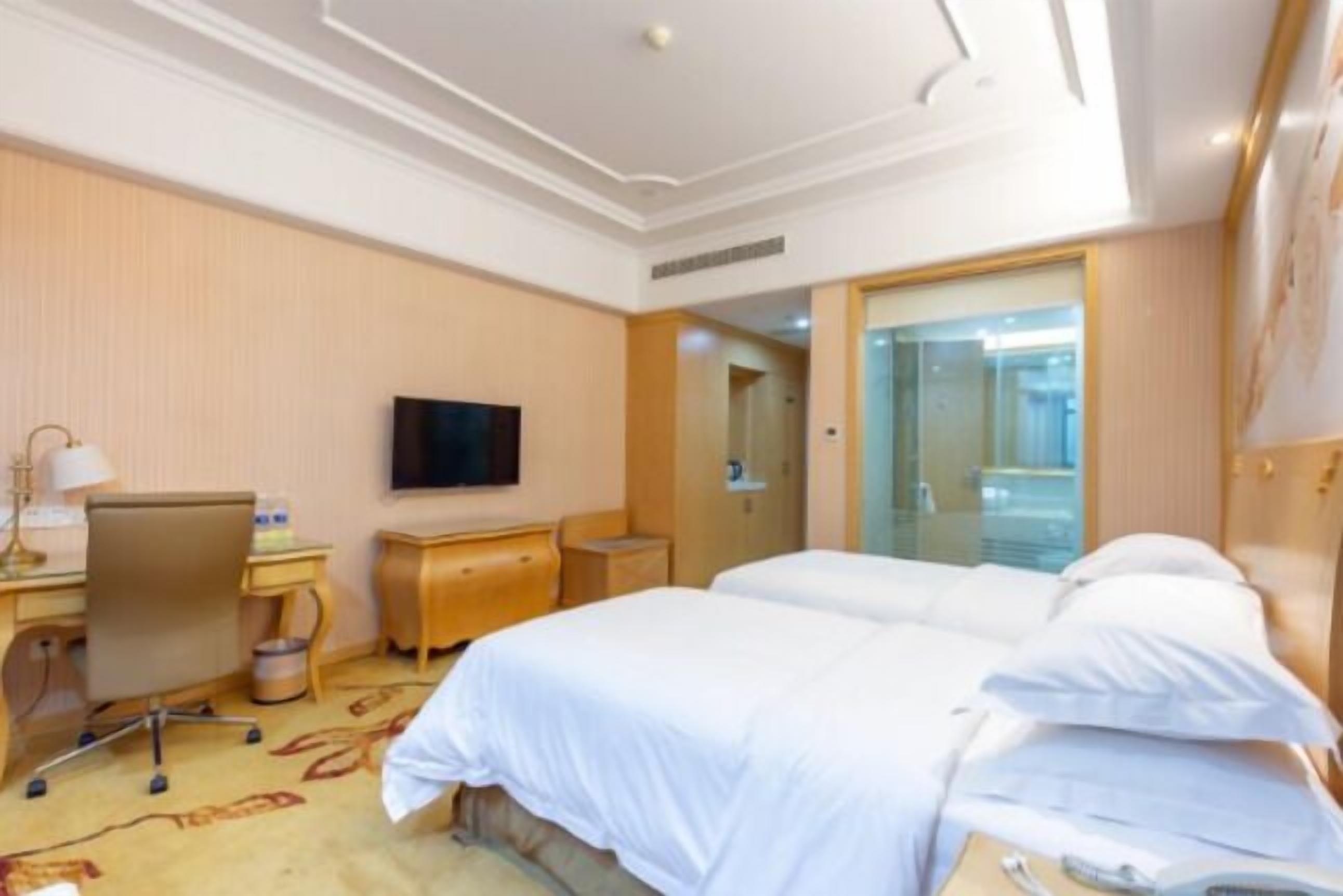 A hotel room with two beds, a desk with a chair, a television, and a bathroom visible through a glass door.