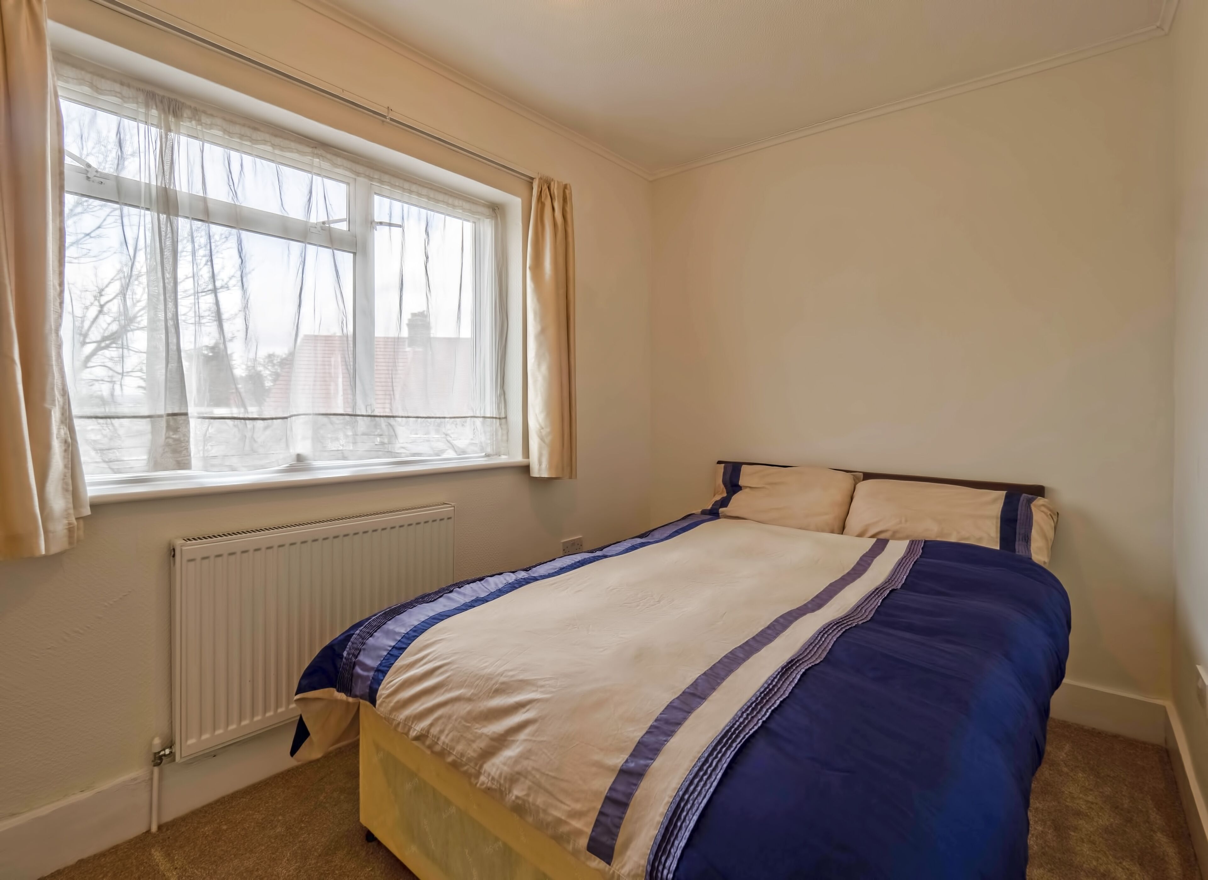 A bedroom with a bed, a window with curtains, a radiator, and a carpeted floor.