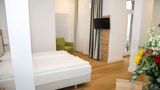 A modern hotel room with a bed, a green armchair, a TV mounted on the wall, and a wooden floor. - Perkeo Apartments: Studio 3 | Heidelberg-Altstadt (Heidelberg)