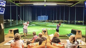 Una sala de simulación de golf con una pantalla grande que muestra un campo de golf, personas jugando y un marcador.