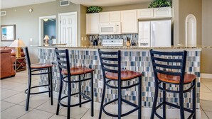 Une cuisine avec un mur recouvert de carreaux, des meubles blancs, un réfrigérateur et un coin bar avec des tabourets.