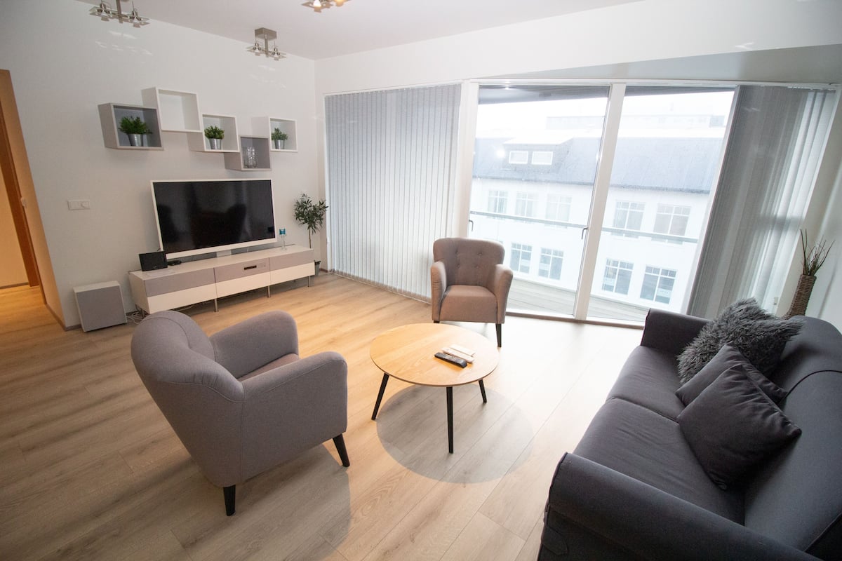 Uma sala de estar moderna com televisão de tela plana, dois sofás, uma mesa de centro e uma grande janela com cortinas.