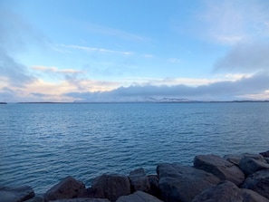 Une mer calme, avec une côte rocheuse et, au loin, des montagnes aux sommets enneigés, sous un ciel partiellement nuageux.