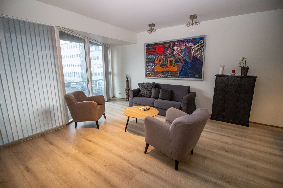 Uma sala de estar moderna com um sofá cinza, duas poltronas, uma mesa de centro de madeira e um quadro na parede.