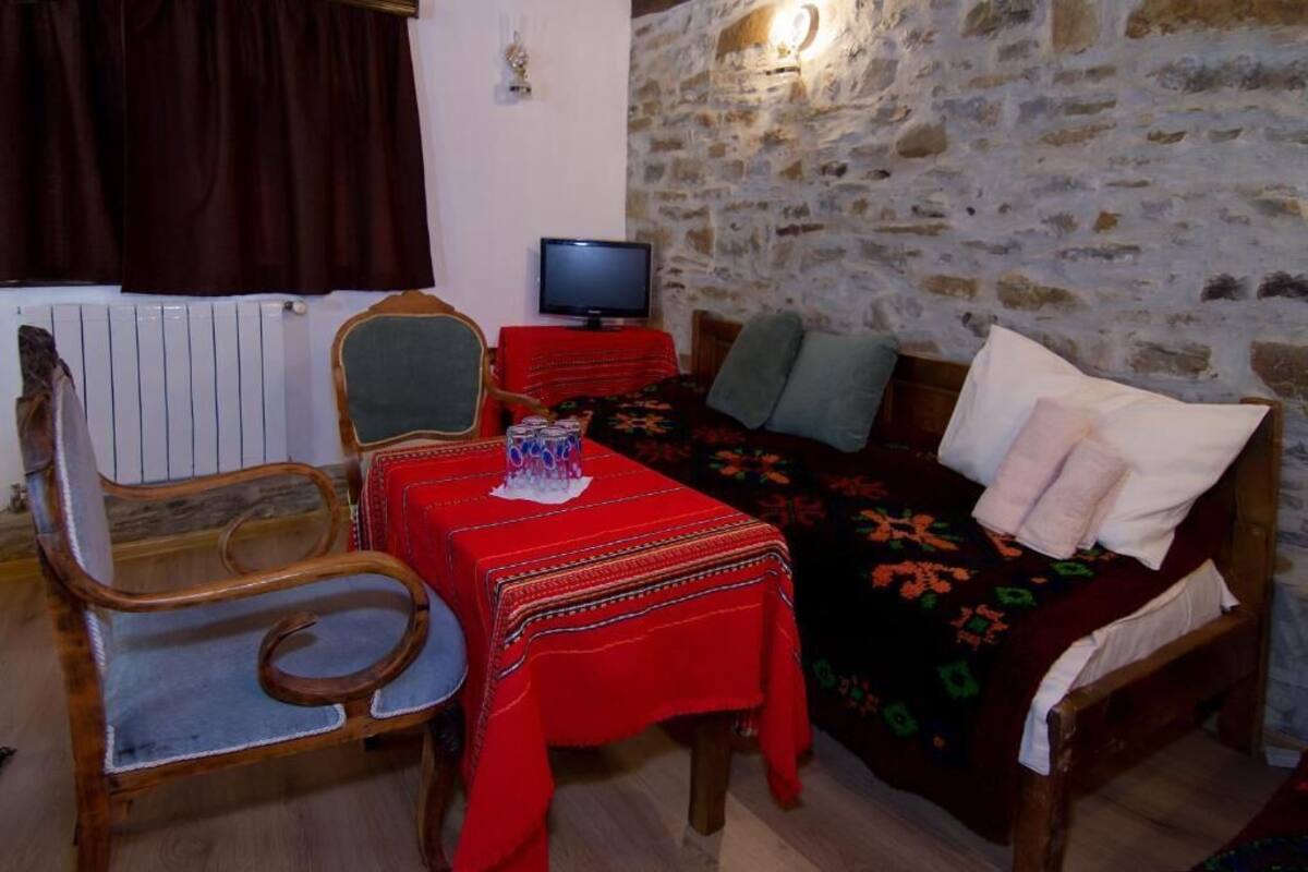Un cuarto acogedor con pared de piedra, una cama de madera con almohadas, una mesa roja con un adorno, una silla de madera y un televisor sobre una mesita.