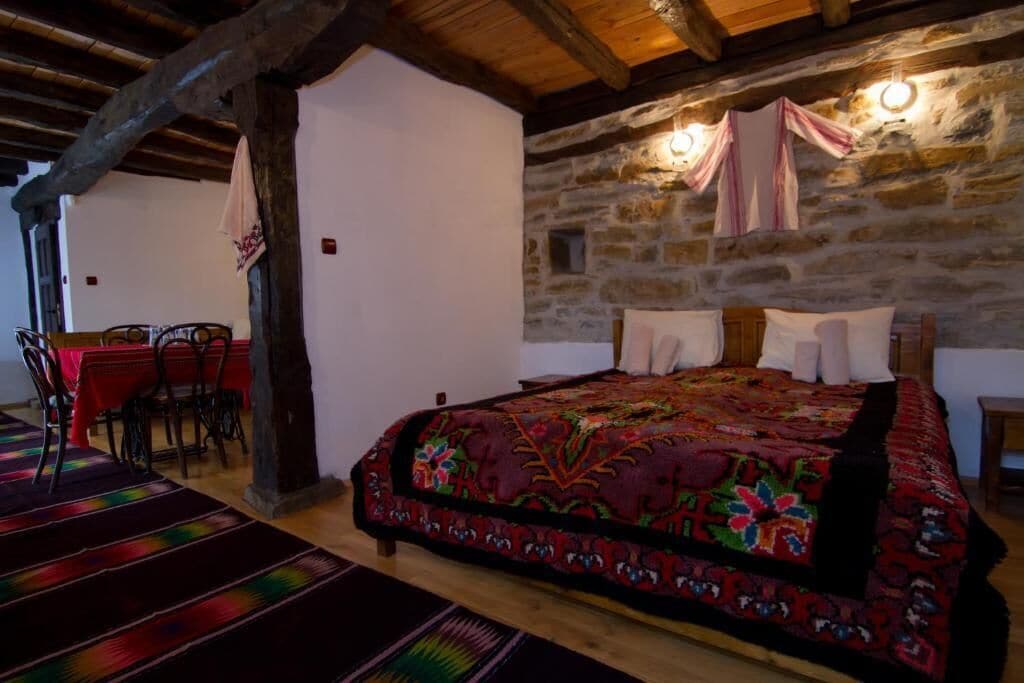 A rustic bedroom with a stone wall, a wooden ceiling, a large bed with a colorful blanket, a dining area with a red tablecloth, and a chair.