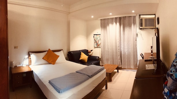 A hotel room with a bed, a desk, a chair, a nightstand, a lamp, a window with curtains, an air conditioning unit, and a wall decoration. - Jeanette Home (New Taipei City)