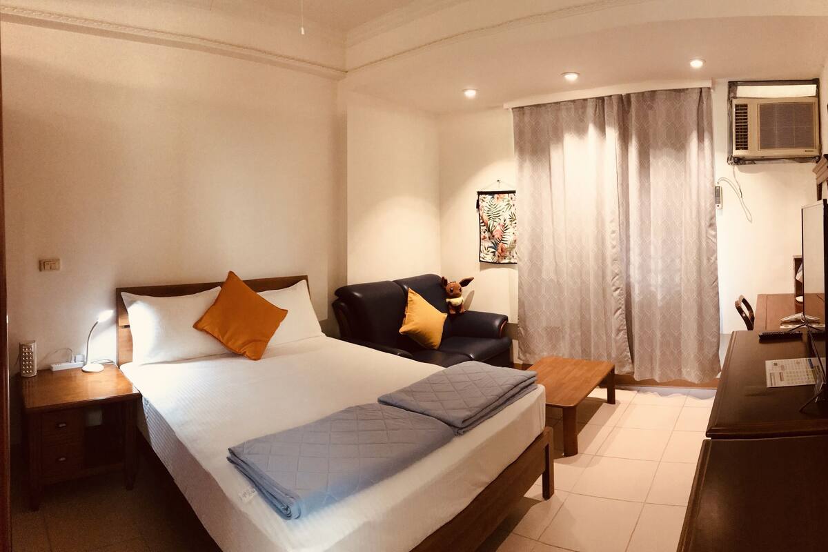 A hotel room with a bed, a desk, a chair, a nightstand, a lamp, a window with curtains, an air conditioning unit, and a wall decoration.