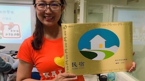A person holding a book with a house and green landscape on the cover, standing in a room with a bed and a desk with various items. - Jeanette Home (New Taipei City)