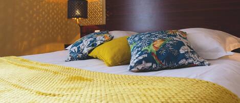 A neatly made bed with a yellow blanket and floral pillows, a dark wooden headboard, and a wall-mounted lamp.
