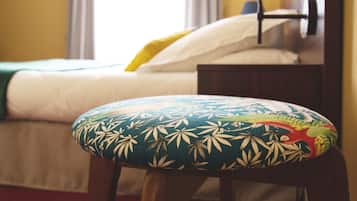 Un lit avec un drap blanc et un oreiller jaune, une tête de lit en bois et un coussin coloré orné d’un motif de feuilles.