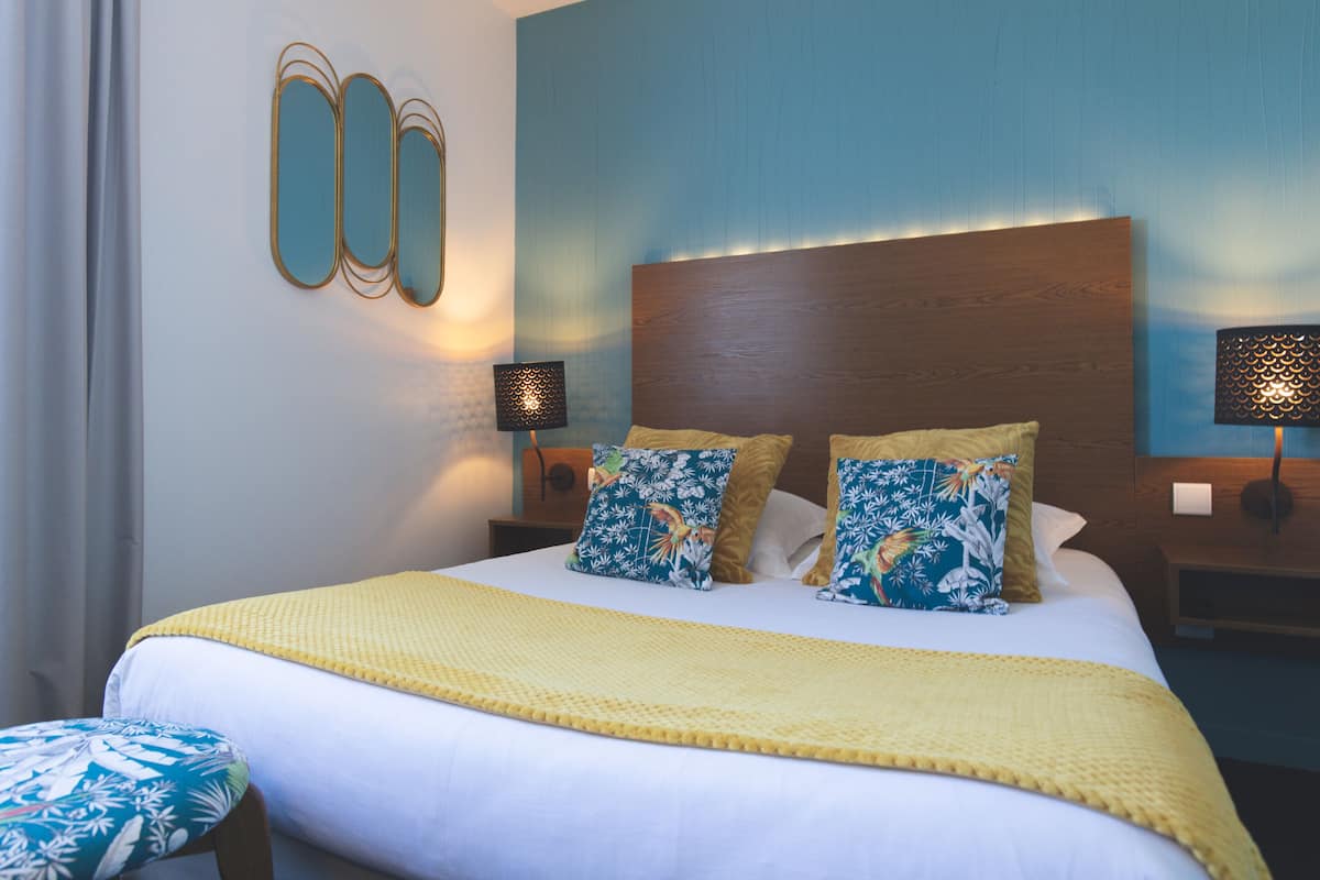 Un lit bien fait, recouvert d’une couverture jaune, avec deux coussins à motifs floraux et un coussin assorti posé sur une tête de lit en bois.