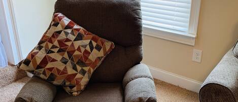 Un sillón marrón con un cojín estampado en una habitación con alfombra color beige, una planta en maceta y una ventana con persianas.