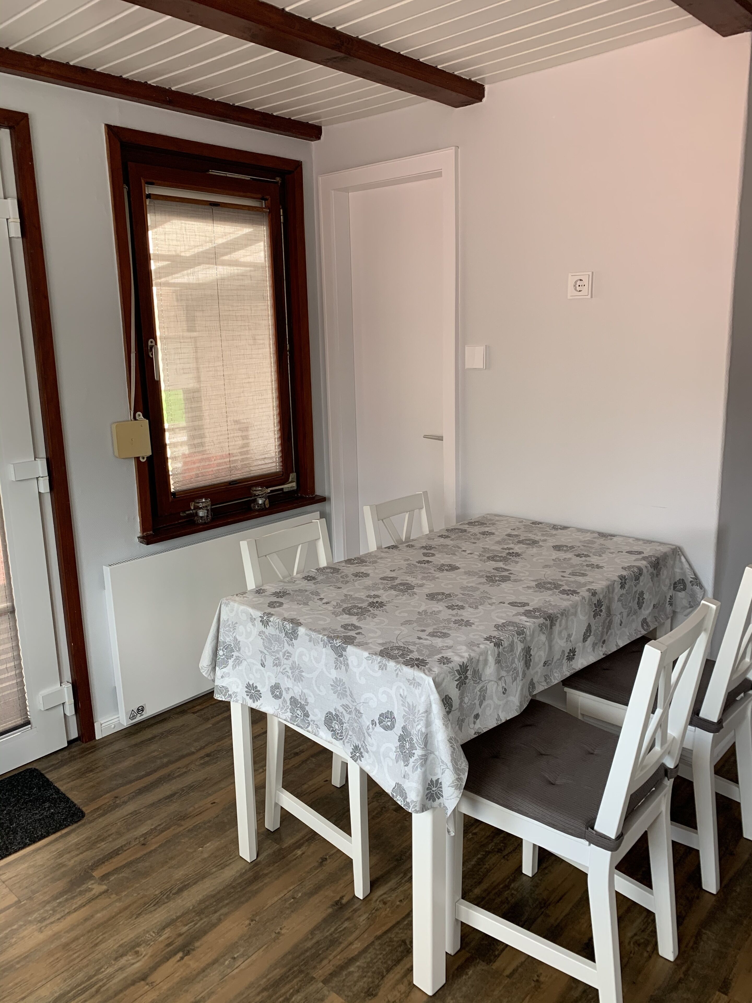 A dining area with a table set for four, white chairs, a window with blinds, and a white door.