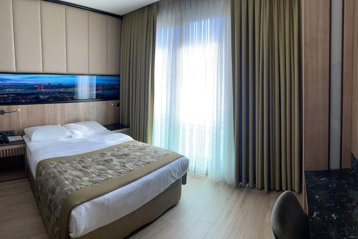Habitación de hotel con cama, televisor, escritorio y un amplio ventanal con cortinas.