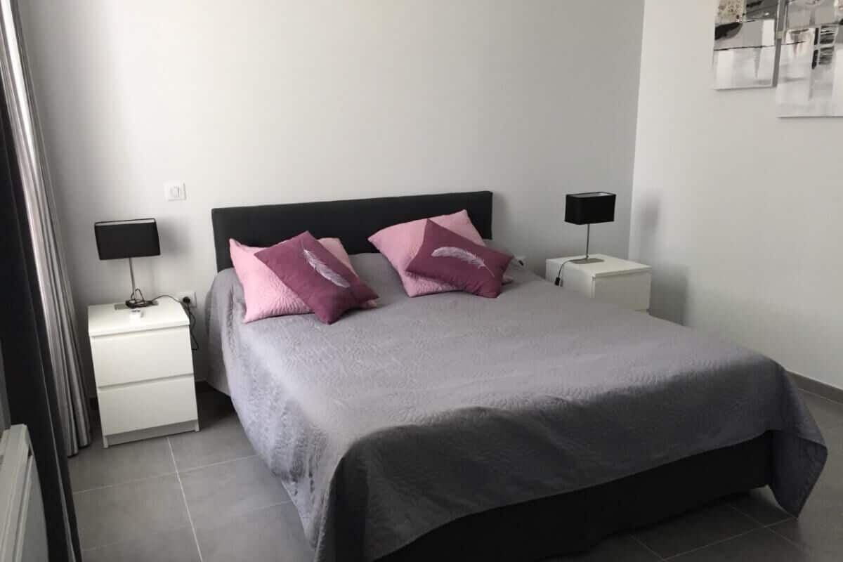 Een net opgemaakt bed met een grijze plaid en twee roze kussens, aan weerszijden met witte nachtkastjes en zwarte lampen.