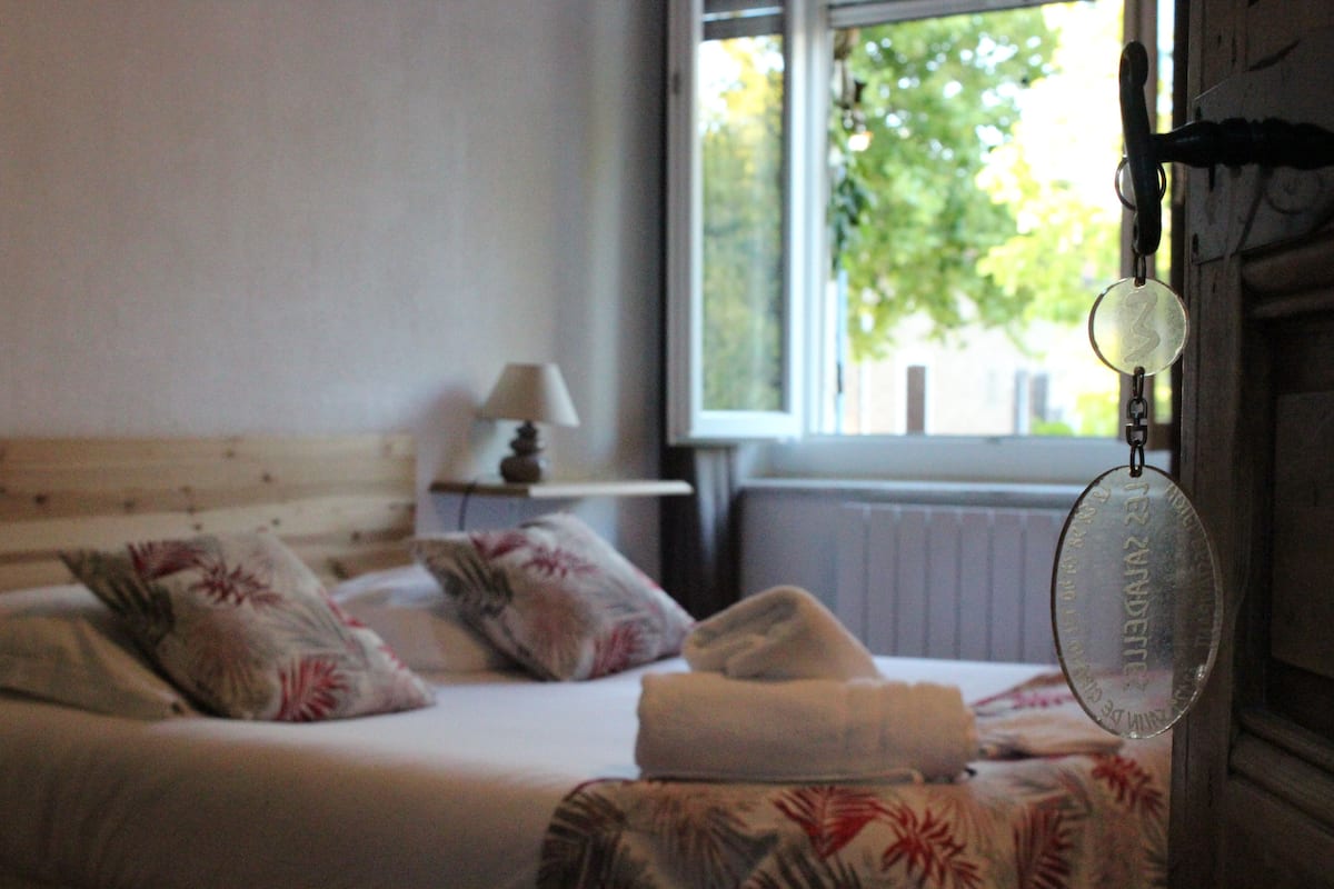 Een slaapkamer met een bed, kussens, een lamp en een raam met uitzicht op bomen.