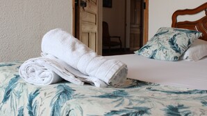 Ein ordentlich bezogenes Bett mit einer blumigen Tagesdecke und einem aufgerollten weißen Handtuch darauf.