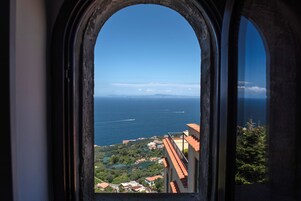 A view through a window with an arched frame, overlooking a coastal area with buildings and greenery.