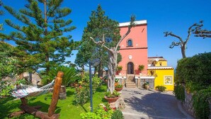 A garden with a hammock, a pathway leading to a building with a red and yellow facade, and a clear blue sky. - Torre Elisabetta (Massa Lubrense)