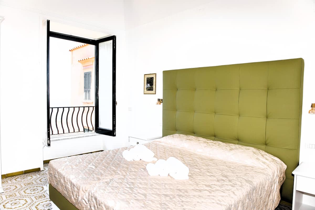A hotel room with a green upholstered headboard, a patterned bedspread, and a window with a black frame.