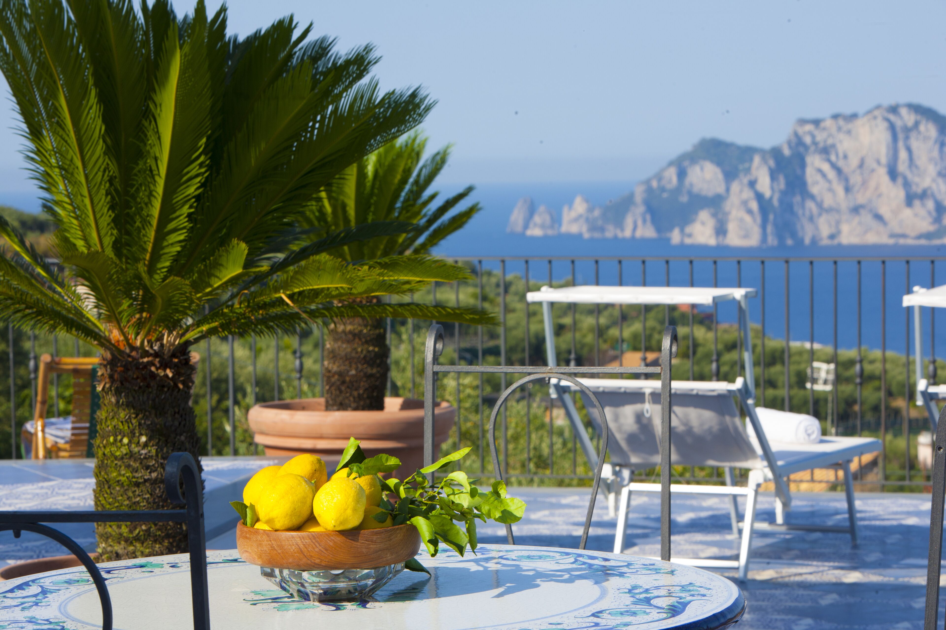 Une table avec un saladier rempli de citrons et une plante en pot, des chaises d’extérieur, et une vue sur la mer et les montagnes.