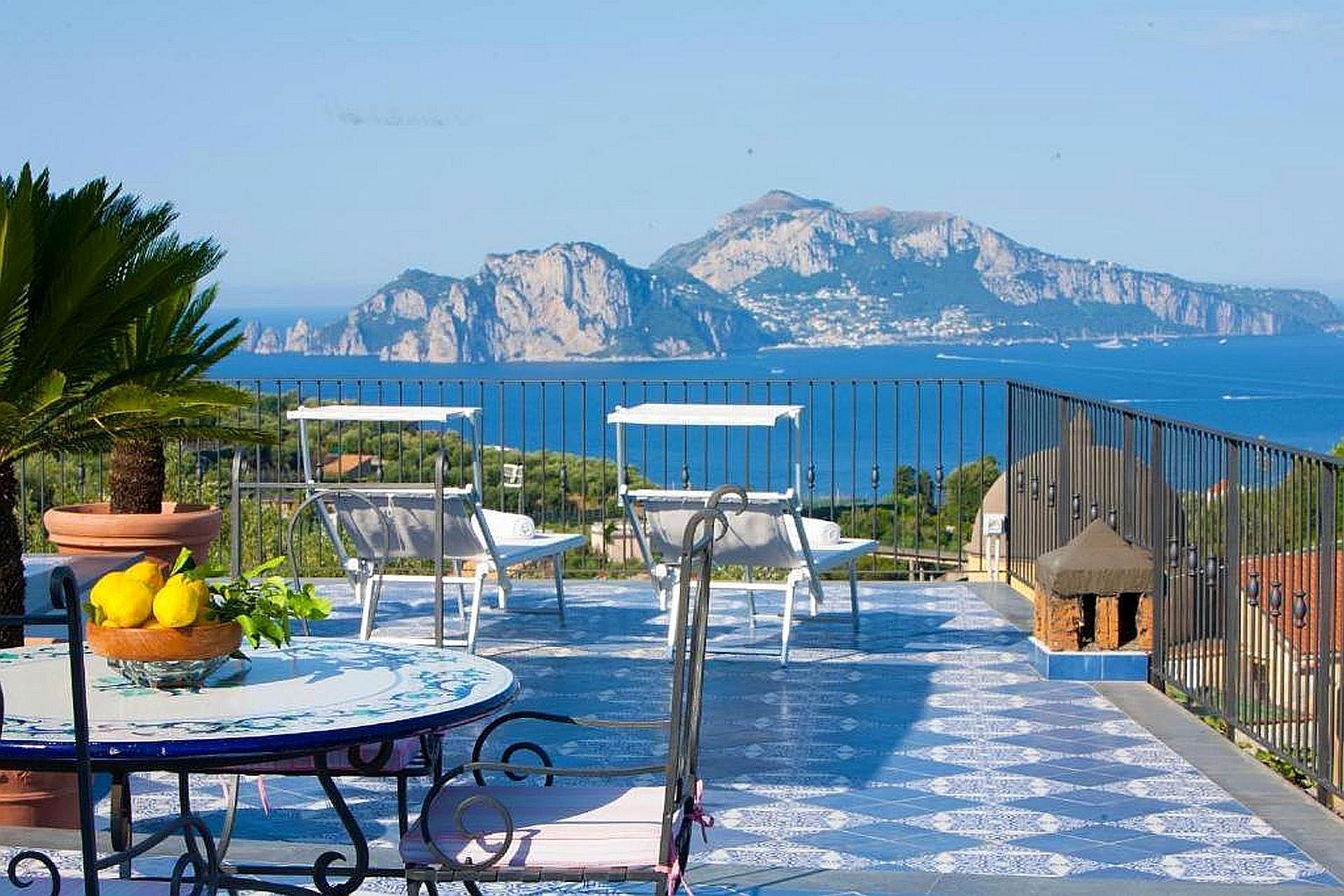 A terrace with a view of the sea and mountains, featuring a table with chairs and a potted plant.