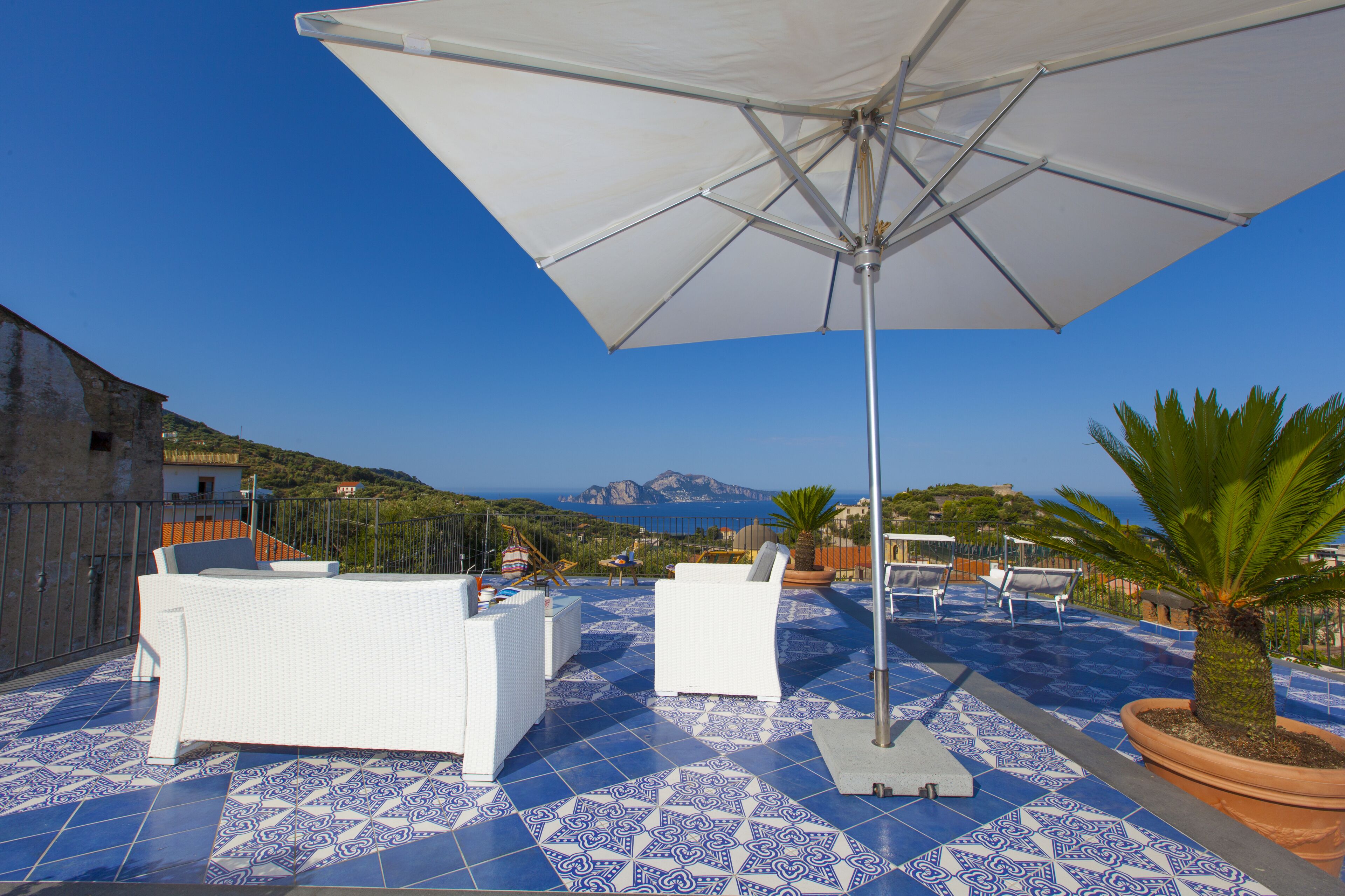 A rooftop terrace with white furniture, a large umbrella, and a view of the sea and mountains.