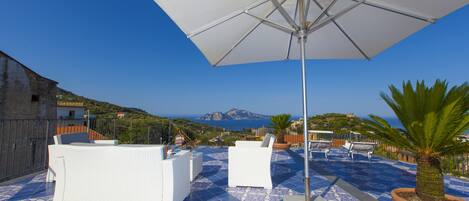 A rooftop terrace with white furniture, a large umbrella, and a view of the sea and mountains.