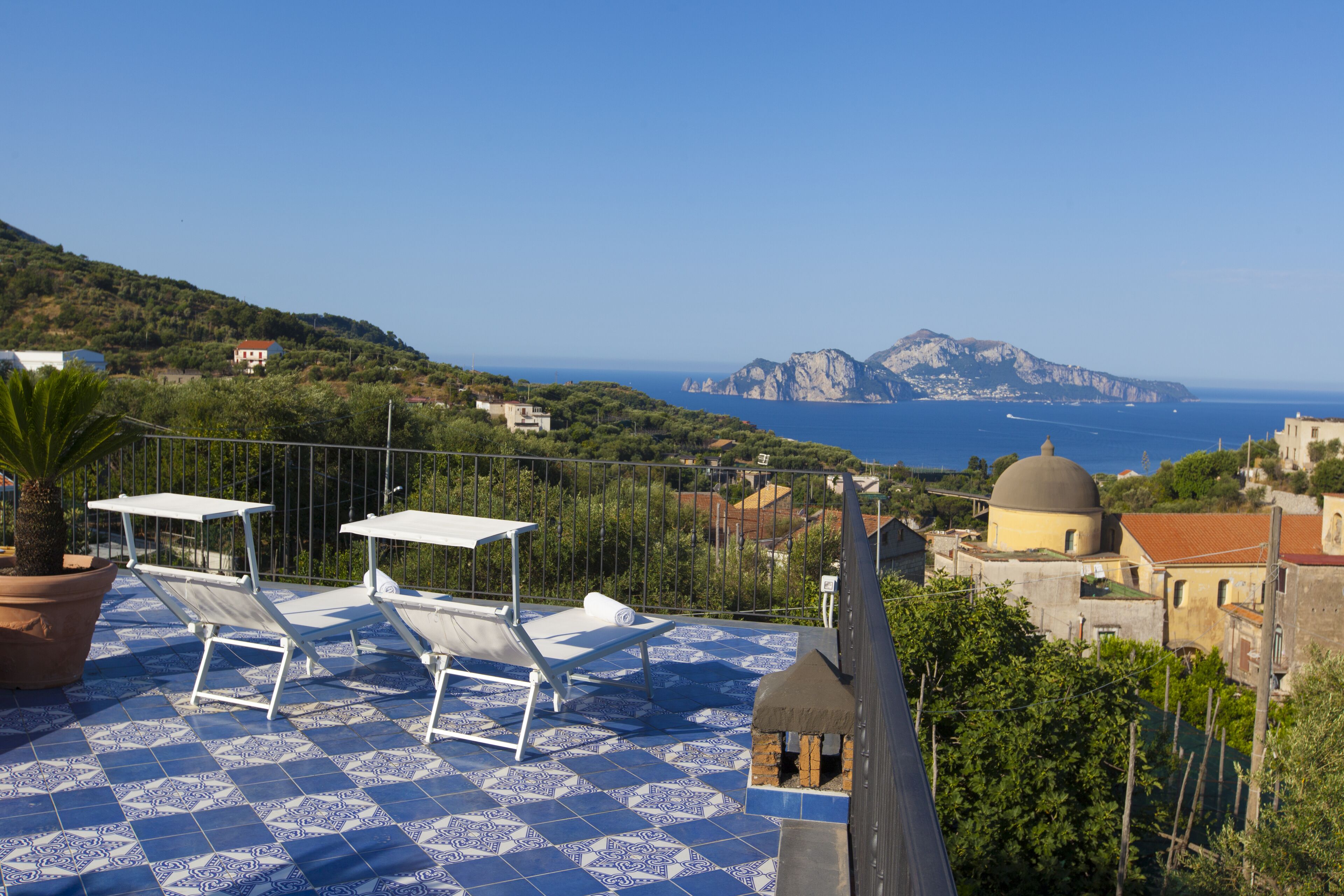 A terrace with sun loungers and a potted plant, overlooking a coastal landscape with buildings and a clear sky.