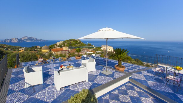 A rooftop terrace with blue patterned flooring, white outdoor furniture, and a large white umbrella. - Torre Elisabetta (Massa Lubrense)