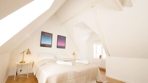 A spacious attic bedroom with a sloped ceiling, a bed with white bedding, two wall-mounted picture frames, a bedside table with a lamp, and a small window with a plant. - Rent A Place 4, Copenhagen (København)