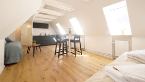 A compact, modern apartment with a loft-style ceiling, a kitchenette, a dining area with a table and chairs, and a large window providing natural light. - Rent A Place 4, Copenhagen (København)
