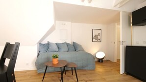 A small, well-lit room with a blue sofa, a small table with a plant, and a wall-mounted light. - Rent A Place 4, Copenhagen (København)