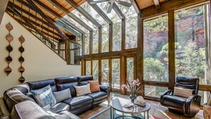 Ein geräumiges Wohnzimmer mit einer ledernen Eckcouch, einem runden Couchtisch und einem großen Fenster mit Blick auf eine bewaldete Landschaft.