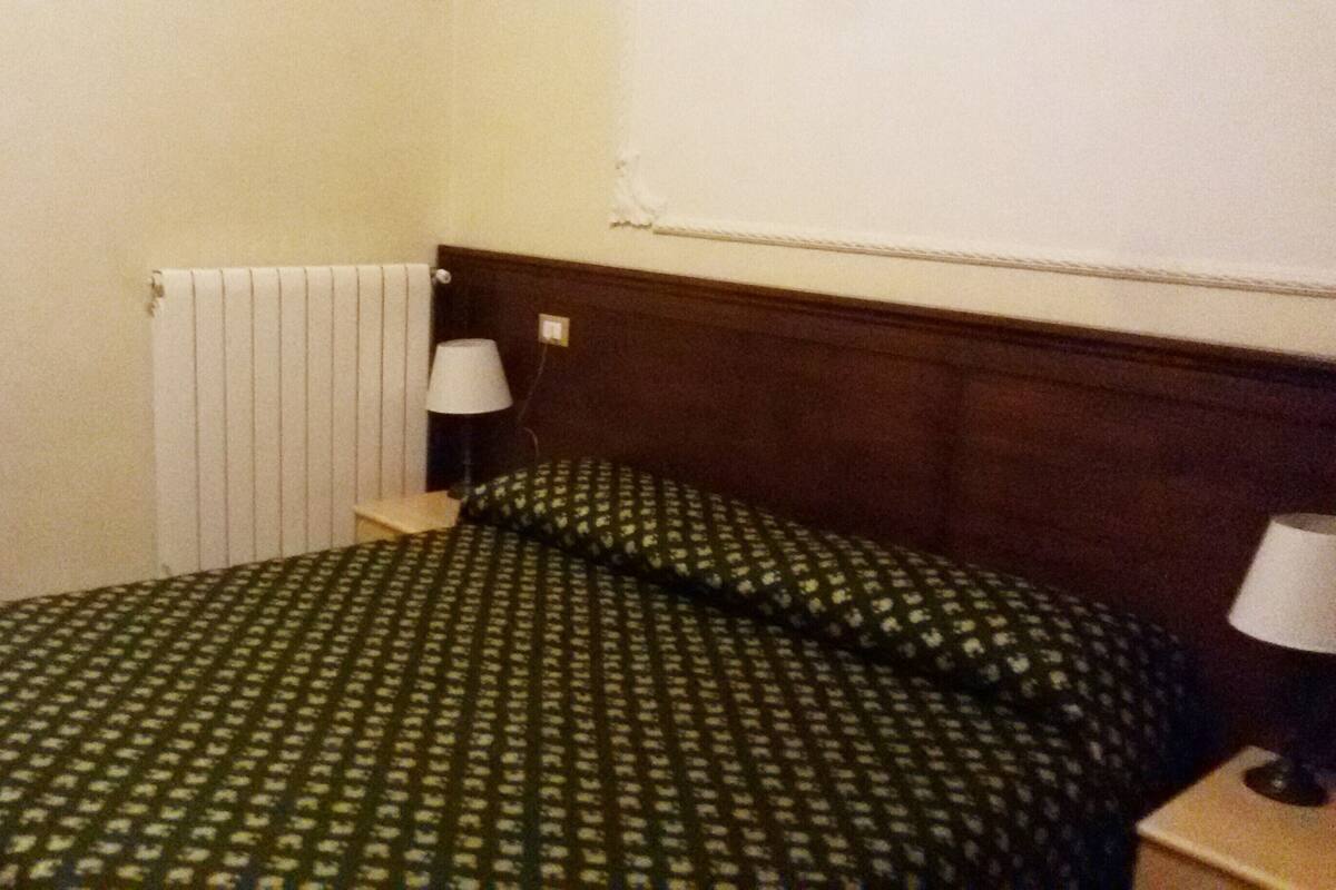 A small, simple room with a bed, a nightstand, a lamp, and a radiator.
