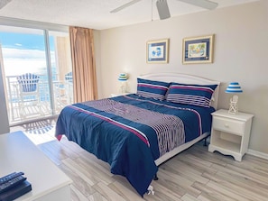 A bedroom with a large bed, a nightstand, a desk, and a view of the outdoors.