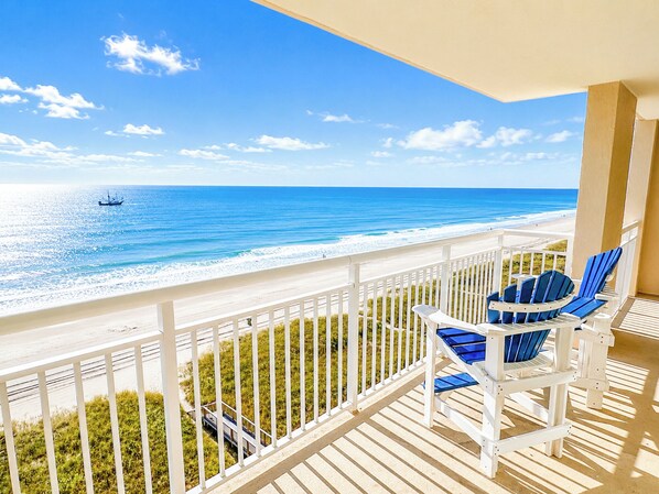 A balcony with a view of the beach and ocean, white railings, and blue chairs.